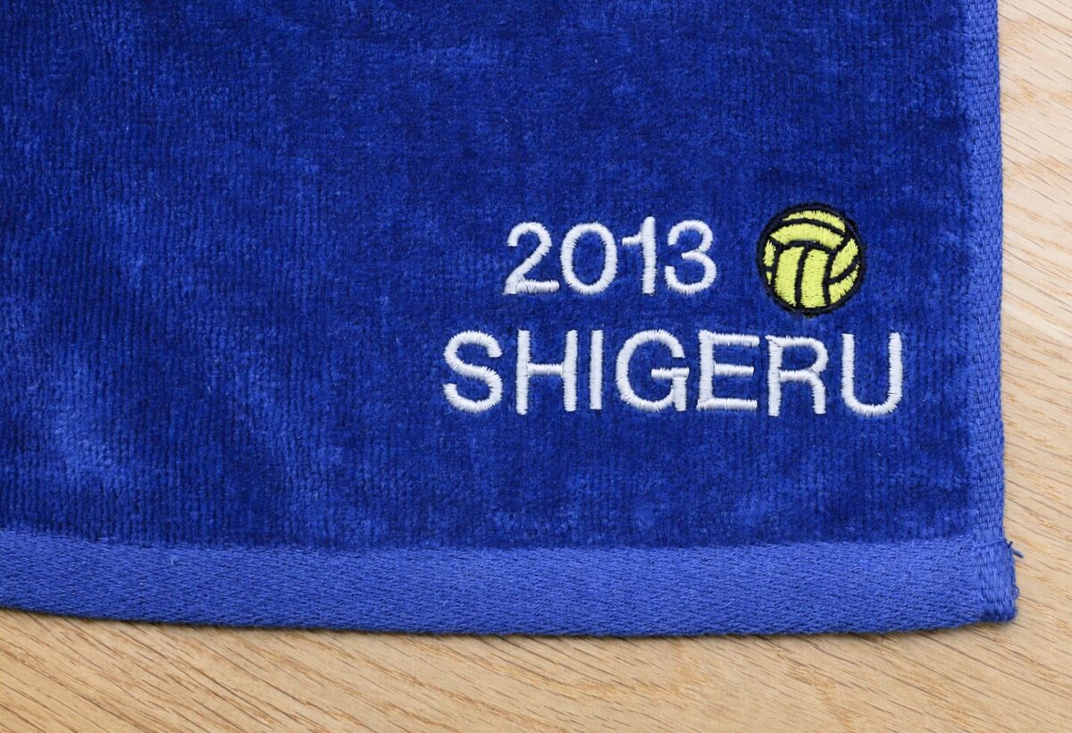 ネイビーブルー色のマフラータオルに、白糸で「2013 SHIGERU」の文字と黄色のバレーボールマークを刺繍した制作例