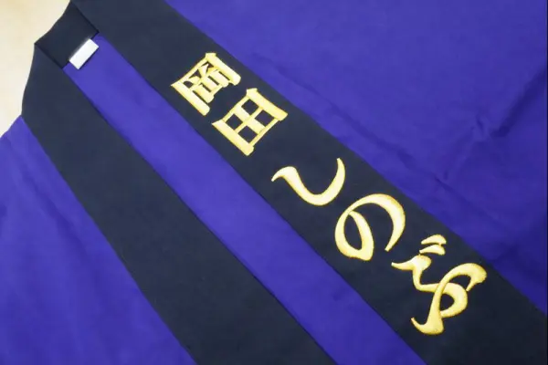 紫地の法被の襟部分に、金糸で「岡田 しのぶ」と名入れ刺繍を施したオーダー刺繍の制作事例