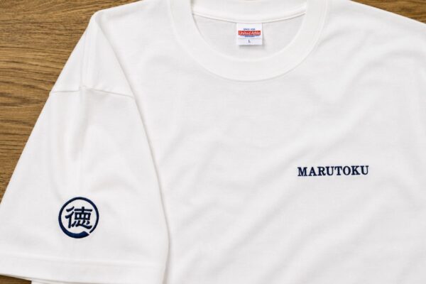 大田区下丸子の丸徳商店様からご注文いただいた白Tシャツ。胸元に「MARUTOKU」、左袖に家紋風の「徳」マークをネイビー糸で刺繍したオリジナルユニフォーム制作事例